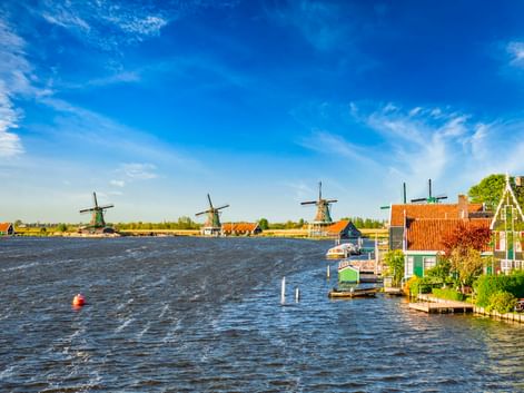Drei historische Windmühlen entlang eines Wasserwegs in Zaanse Schans mit traditionellen niederländischen Häusern mit orangefarbenen Dächern unter blauem Himmel.