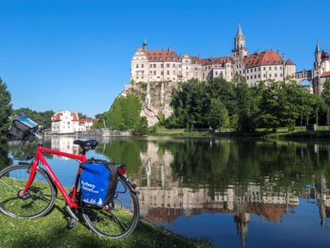 Rotes Fahrrad mit blauen Taschen auf Wiese an der Donau. Schloss Sigmaringen thront auf einem Felsen über dem Wasser und spiegelt sich in der ruhigen Oberfläche.