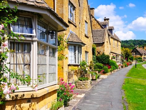 Reihe honigfarbener Steinhäuser mit blühenden Gärten entlang einer Dorfstraße in den Cotswolds unter blauem Himmel mit weißen Wolken.