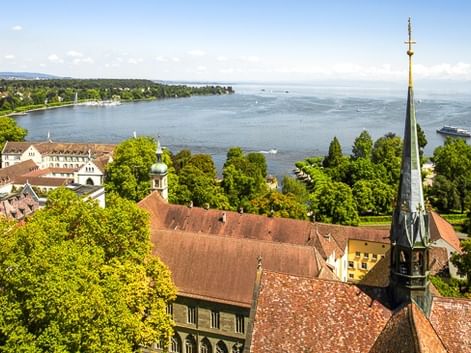 Rote Ziegeldächer und Kirchturm mit Blick auf den Bodensee mit grünen Bäumen, Segelbooten auf dem Wasser und fernem Ufer unter blauem Himmel.