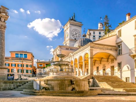 Historischer Platz in Udine mit verziertem Brunnen, weißem Uhrenturm mit blauer Kuppel, Arkadenloggia und bunten Gebäuden unter blauem Himmel.