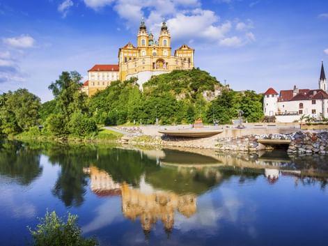 Gelbes barockes Stift Melk auf grünem Hügel, gespiegelt in der ruhigen Donau, mit weißer Kirche am rechten Ufer sichtbar.