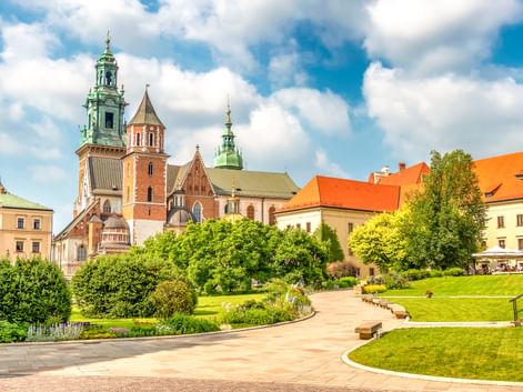 Wawel-Kathedrale in Krakau mit markanten grünen Kupfertürmen und roten Backsteinmauern, umgeben von gepflegten Rasenflächen und Gärten unter blauem Himmel.