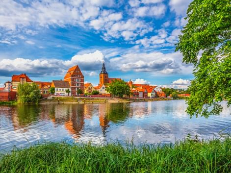 Blick auf Havelberg über den Fluss mit historischen Gebäuden mit roten Dächern und Kirchturm, die sich im ruhigen Wasser spiegeln.