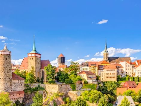 Panoramablick auf Bautzens Altstadt mit mittelalterlichen Türmen, Kirchtürmen und bunten Gebäuden auf einem Felsenhügel unter blauem Himmel.
