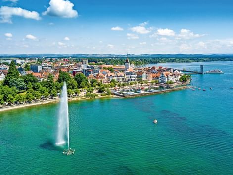 Luftaufnahme von Friedrichshafen am Bodensee mit hohem Springbrunnen im türkisfarbenen Wasser, historischer Altstadt, Kirchturm und Fahrgastschiff.