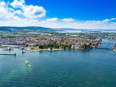Luftaufnahme von Konstanz und Kreuzlingen am Bodensee mit historischen Gebäuden, einem Hafen und Booten auf türkisfarbenem Wasser unter blauem Himmel.