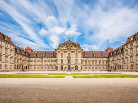 Symmetrisches Barockschloss mit roten Ziegeldächern und Sandsteinfassade um einen Innenhof mit grünem Rasen unter dramatischem Wolkenhimmel.
