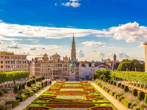 Formale Gärten am Mont des Arts in Brüssel mit bunten geometrischen Blumenbeeten, gestutzten Hecken und Stadtsilhouette mit Kirchturm unter blauem Himmel.