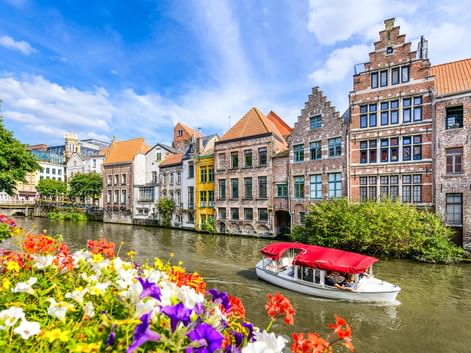 Touristenboot mit rotem Dach auf Kanal in Gent, gesäumt von bunten historischen Gebäuden. Bunte Blumen rahmen den Vordergrund.