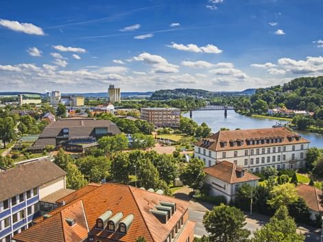Aerial view of Hameln with red-tiled roofs, the Weser River flowing through, a bridge, and green hills under a blue sky with white clouds.