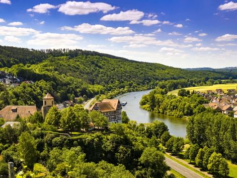 Luftaufnahme der Burg Homburg mit Turm neben einem Fluss, umgeben von bewaldeten Hügeln, grünen Wiesen und Dorfhäusern unter blauem Himmel.