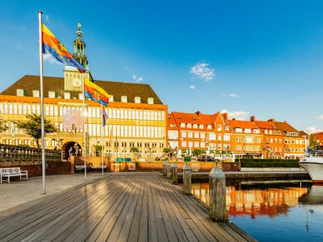 Emder Rathaus mit gelb-weißer Fassade und Turm hinter einer hölzernen Hafenpromenade mit Flaggen. Orangefarbene Backsteingebäude und Boote im Hafen.