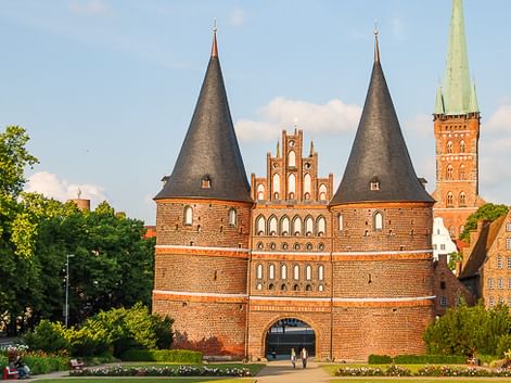 Das Holstentor, ein mittelalterliches Stadttor aus rotem Backstein in Lübeck mit zwei runden Türmen und Kegeldächern, umgeben von Grünflächen.
