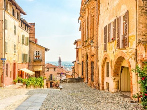 Kopfsteinpflasterstraße in Saluzzo mit ockerfarbenen historischen Gebäuden, Fensterläden und einem Kirchturm in der Ferne.