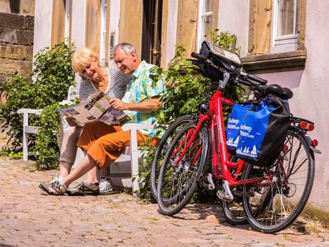 Zwei Radfahrer sitzen auf einer weißen Bank und lesen eine Karte in Bad Wimpfen. Zwei rote Tourenräder mit blauen Taschen stehen daneben.