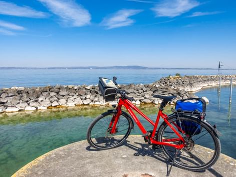 Rotes Tourenrad mit blauen Taschen auf steinerner Mole am Bodensee. Steinerner Wellenbrecher und ruhiges blaues Wasser unter klarem Himmel.