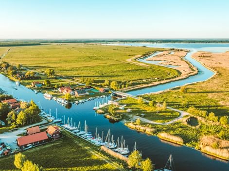 Luftaufnahme des Memeldeltas mit gewundenen Wasserwegen, grünen Feldern, Yachthafen mit Segelbooten und kleinem Dorf mit roten Dächern.