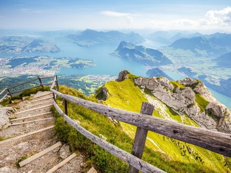 Bergweg mit Holzzaun auf dem Pilatus mit Blick auf den Vierwaldstättersee. Grüne Almwiesen, Felsgipfel und blauer See mit umliegenden Bergen.