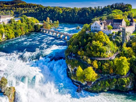 Luftaufnahme des Rheinfalls bei Schaffhausen mit kraftvollem Wasserfall, historischem Schloss, Steinbrücke und grünen Wäldern unter blauem Himmel.