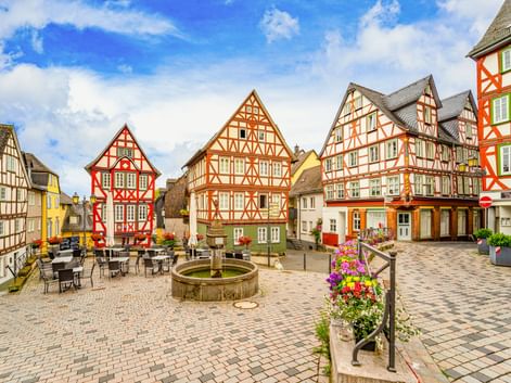 Domplatz square in Wetzlar with colorful half-timbered houses, a stone fountain, outdoor café seating, and flower-decorated benches under blue sky.
