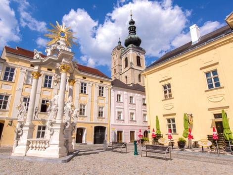 Baroque square in Krems-Stein with ornate white monument, pastel buildings, and church tower under blue sky with white clouds.