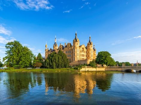 Schweriner Schloss, ein prächtiger Palast mit goldenen Kuppeln und Türmen, liegt auf einer Insel umgeben von ruhigem Seewasser unter blauem Himmel.