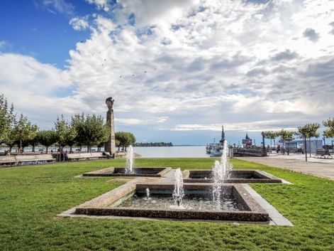 Stufenbrunnen mit Wasserfontänen auf grüner Wiese am Hafen in Konstanz. Bodensee und historisches Hafengebäude im Hintergrund unter bewölktem Himmel.