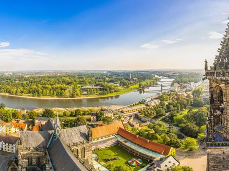 Luftaufnahme von Magdeburg mit gotischem Domturm im Vordergrund, Elbe schlängelt sich durch grüne Landschaft, Stadtgebäude unter blauem Himmel.