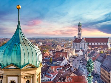 Luftaufnahme von Augsburg mit grünen Zwiebeltürmen im Vordergrund und dem hohen Rathausturm über der historischen Altstadt bei Sonnenuntergang.