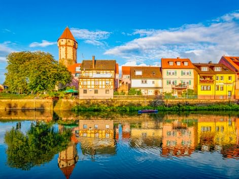Spitzer Turm, ein runder Turm mit orangefarbenem Dach in Wertheim, spiegelt sich im Main. Bunte Fachwerkhäuser säumen das Flussufer.