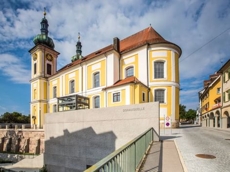 Gelb-weiße Barockkirche mit grünen Kuppeltürmen in Donaueschingen. Steinmonument mit Aufschrift 'Donauquelle' im Vordergrund.