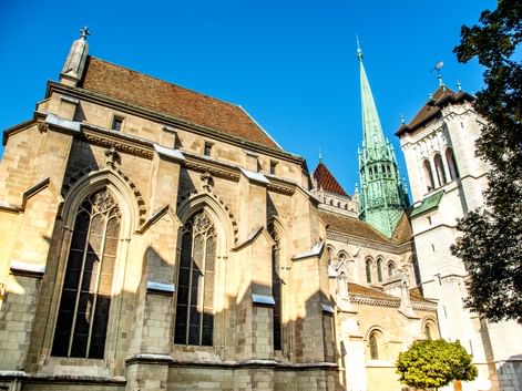 St. Peter's Cathedral in Geneva with Gothic arched windows, stone facade, and distinctive green copper spire against a clear blue sky.