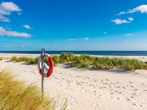 Weißer Sandstrand in Dueodde auf Bornholm mit rot-weißem Rettungsring an Stange, Dünen mit Strandgras und blauem Meer unter klarem Himmel.
