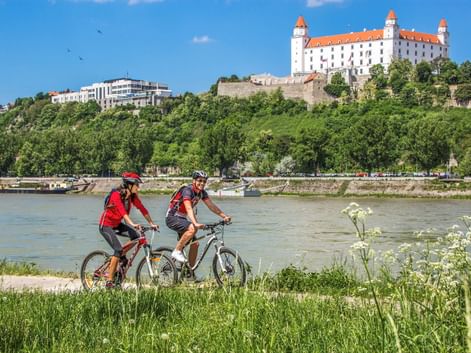 Zwei Radfahrer fahren entlang der Donau mit der Burg Bratislava auf dem Hügel. Weiße Blumen blühen im Vordergrund.