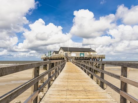 Langer Holzsteg führt zu einem Pfahlbau am Strand von St. Peter-Ording. Breiter Sandstrand und dramatischer Wolkenhimmel im Hintergrund.