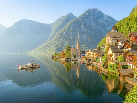 Hallstätter See mit traditionellem Alpendorf am Ufer, Kirchturm, Bergen und einem Boot auf ruhigem Wasser, das die Szenerie spiegelt.
