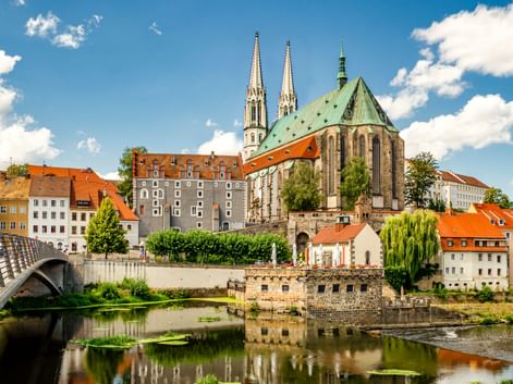 St. Peter and Paul Church with twin spires and green copper roof in Görlitz, reflected in the Neisse River with historic buildings and bridge.