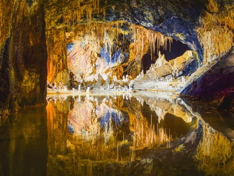 Beleuchtete Feengrotte Saalfeld mit farbigen Stalaktiten und Stalagmiten, die sich in stillem unterirdischem Wasser spiegeln, mit goldener und blauer Beleuchtung.