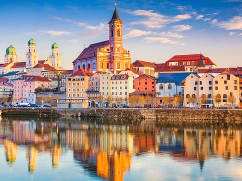 Passau old town skyline with colorful buildings along the Danube riverfront. Church towers with green domes and a yellow church are reflected in the water.