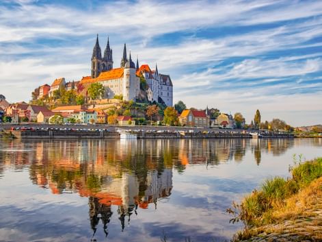 Historisches Meißen mit Albrechtsburg und Dom auf dem Burgberg über der Elbe. Bunte Häuser säumen das Ufer, perfekt im ruhigen Wasser gespiegelt.