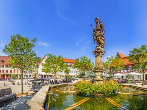 Historischer Brunnen mit Statue auf Säule auf dem Öhringer Marktplatz, umgeben von bunten Blumen, Bäumen, Bänken und traditionellen Gebäuden.