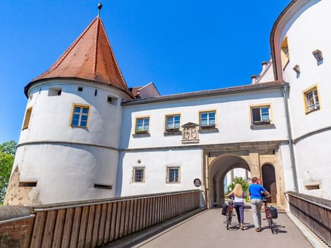 Two cyclists walking their bikes across a bridge toward Wörth Castle entrance. White castle walls with red-tiled tower under blue sky.