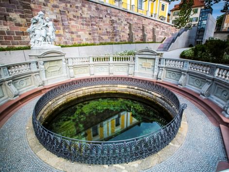 Rundes Steinbecken mit verziertem Eisengeländer markiert die Donauquelle in Donaueschingen, umgeben von weißen Balustraden und einer Statue.