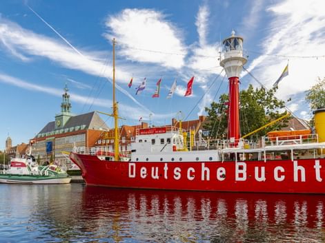 Rotes Feuerschiff Deutsche Bucht mit weißem Leuchtturm im Hafen von Emden. Historische Gebäude und blauer Himmel mit Wolken im Hintergrund.