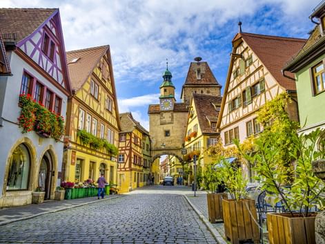 Kopfsteinpflasterstraße in Rothenburger Altstadt mit bunten Fachwerkhäusern und mittelalterlichem Torturm mit Uhr unter blauem Himmel.