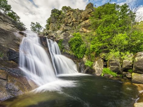 Wasserfall Cascada del Diablo in Villanueva de la Vera, der in ein natürliches Becken fließt, umgeben von üppiger grüner Vegetation und Felsen.