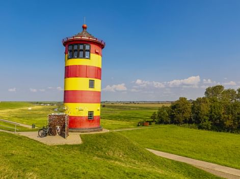 Rot-gelb gestreifter Pilsumer Leuchtturm in Krummhörn auf grüner Wiese mit geparkten Fahrrädern unter blauem Himmel.
