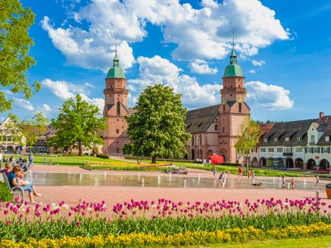Freudenstädter Stadtkirche mit Zwillingstürmen und grünen Kupferdächern mit Blick auf einen Brunnenplatz mit bunten Tulpen und Menschen an einem sonnigen Tag.