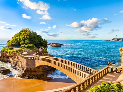 Steinerne Fußgängerbrücke mit Balustraden zu einer grünen Felseninsel in Biarritz. Blaues Meer, Sandstrand und Küstenlandschaft unter Sonnenhimmel.